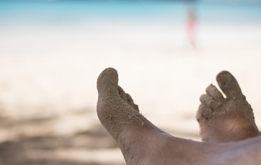 Men's feet in a relaxed position and with sand are on a beautiful sea beach on a sunny day. A blurred person silhouette in a pink swimming suit is visible against the blue sea. Hello, summer holidays!