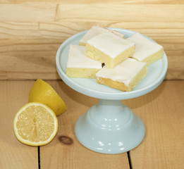 Homemade lemon brownies on a jade green cake plate and two lemon halves against a wood background.