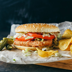 Chiken burgers on wooden tray. Two Homemade hamburgers, pickles and chips on a black metal background. Baking paper