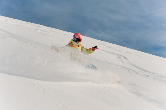 Female snowboarder in colorful sportswear and pink helmet riding down the powder snow hill