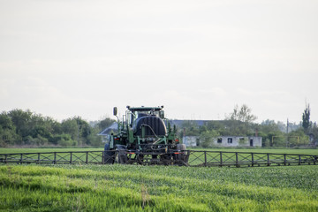 Fototapeta premium Tractor with high wheels is making fertilizer on young wheat. The use of finely dispersed spray chemicals. Tractor with a spray device for finely dispersed fertilizer. Tractor on sunset background.