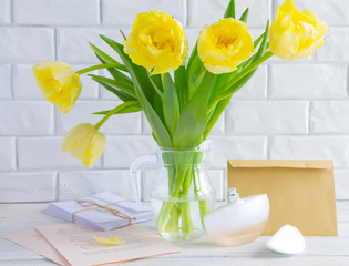 On a background of a white brick wall a vase with yellow tulips, an envelope and a bottle of perfume.