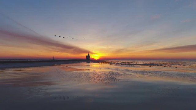 Dramatic sunrise with scenic Lighthouse and flying Canadian Geese, reflected in calm water surface.
