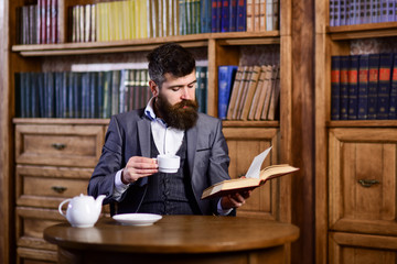 Businessman sits in vintage interior, holds book and cup of tea.