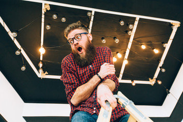 Hipster creative beard guy in eyeglasses balancing on construction worker climbing a ladder while working on home interior over black ceiling background. Anxiety unhappy expression concept. - obrazy, fototapety, plakaty