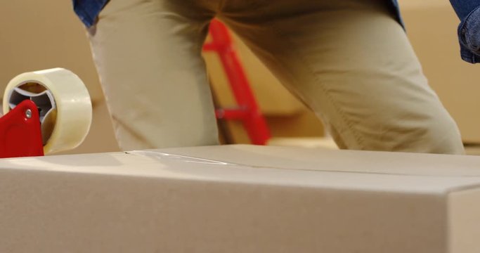 Close up of the man's hands packing a carton box with a sticky tape. Indoors