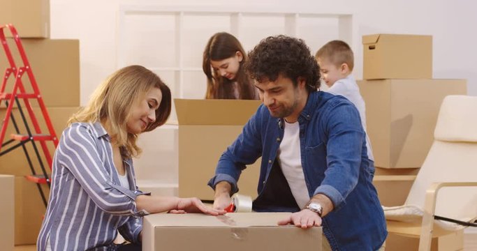 Portrait shot of the wife and husband packing a box with a sticky tape and smiling to the camera, then their children coming to them from the background. Inside