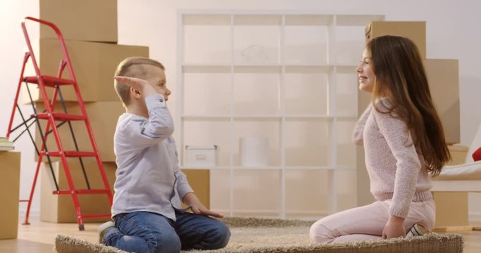 Cute two children untwisting the carpet in the living room among paked boxes after they moving in the new home. Indoor