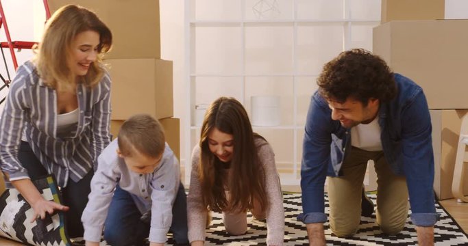 Happy family of parents and two children untwisting the carpet in the living room among paked boxes when they moving in the new home. Indoors