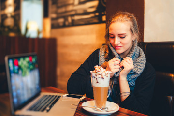A young girl drinks cappuccino with marshmallow working at the laptop sitting in a cozy warm cafe. Morning coffee in internet cafe - Beautiful girl checking news on web and drinking cappuccino. Toned