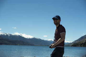 Hombre joven con gorra frente a paisaje de picos de montañas nevados y verdes con cielo azul reflejado en un lago en Nueva Zelanda