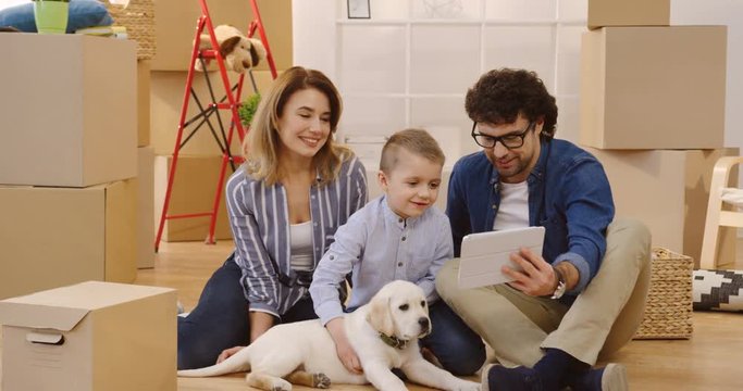Happy mother, father and son sitting on the floor with their puppy among carton boxes and having videochat, waving their hands and talking. Indoors