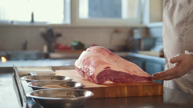 Butcher adding spices into large piece of fresh raw meat lying on a wooden board in a commercial kitchen