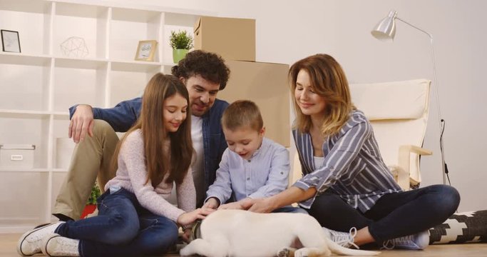Portrait shot of the joyful happy family stroking a cute puppy while sitting on the floor surrounded by unpacked boxes and home stuff while moving in the new apartment. Inside