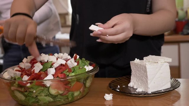 Female hands cook adding cheese to tasty healthy salad prepared in bowl while her friends gathering in the background in kitchen 