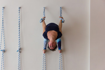 Iyengar yoga student in spider-like posture holding to ropes on white wall background. Young female...