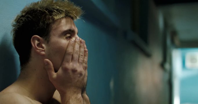 Side view of disappointed and tired sportsman wiping face and leaning on wall in semilit locker room.