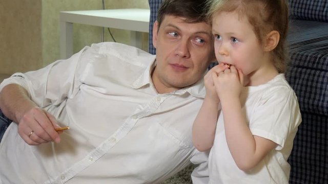 man and daughter watching television, sitting on the floor eating snacks