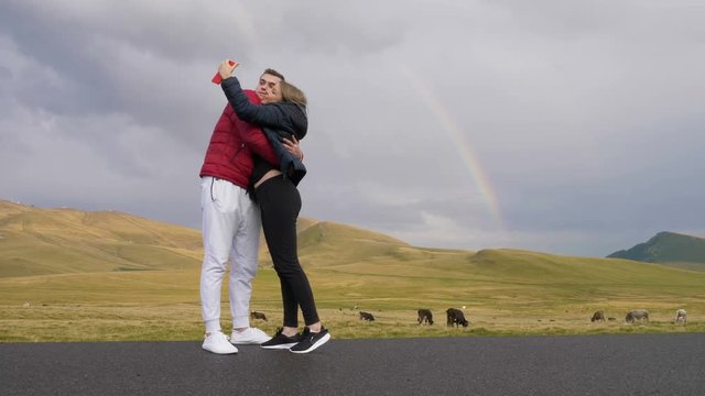 Affectionate teen couple in love hugging and kissing while they taking selfie pics with rainbow and picturesque countryside landscape 