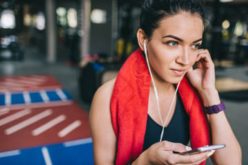 Crlose up portrait of a beautiful smiling and sporty young woman listening to music from her smartphone, having a break after workout in the gym. People, technology, fitness and sport concept.