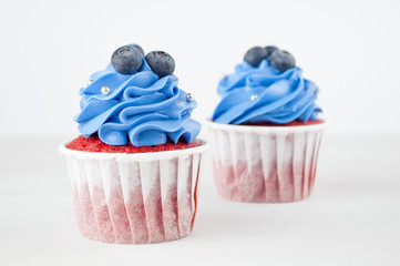 Cupcake red velvet with blue whipped cream decorated with blueberry, silver confectionery balls on white wood table. Picture for a menu or a confectionery catalog. Top view.