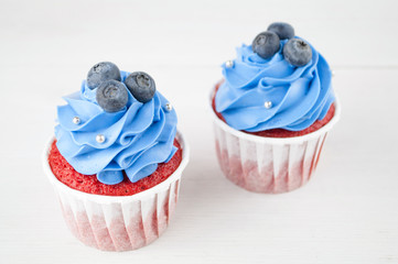 Cupcake red velvet with blue whipped cream decorated with blueberry, silver confectionery balls on white wood table. Picture for a menu or a confectionery catalog. Top view.