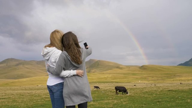 Young playful girls near a pastoral landscape field with cows taking a selfie photo with rainbow in background