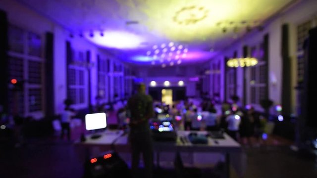 Defocused rear view of music DJ mixing dancing in front his turntables and mixer and laptop during party wedding - illumination with laser light of his back macarena large room with guests