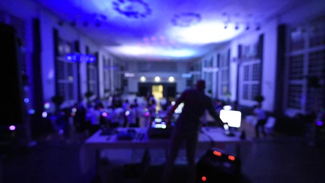 Defocused rear view of music DJ mixing dancing in front his turntables and mixer and laptop during party wedding - illumination with laser light of his back and large room macarena dancing guests