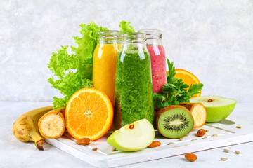 Green, yellow, purple smoothies in currant bottles, parsley, apple, kiwi, orange on a gray table.