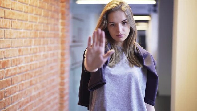 Calm and attractive young woman looking at camera, saying no and stretching her hand to stop somebody off camera while standing in a building corridor with brick walls Handheld slow motion medium shot