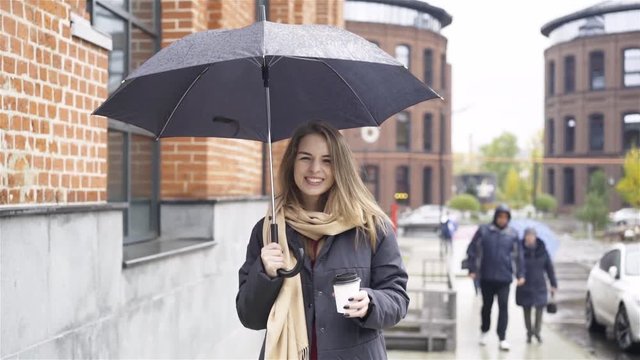 Cheerful blonde businesswoman wearing a coat and a scarf is standing with a coffee under an umbrella in a street. Handheld slow motion medium shot