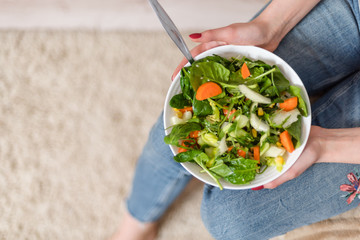Sitting woman hold and eating vegan salad with arugula, celery and spinach in white plate. Healthy eating