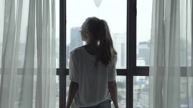 Young woman standing near big window and looking out her apartment on the city buildings in slow motion