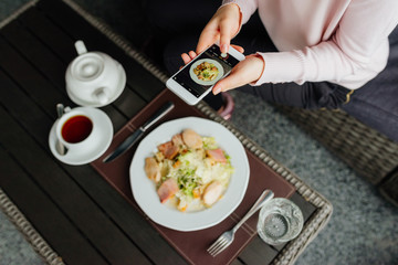 Girl hand holding smartphone and taking photo of food on wooden table. Top view.