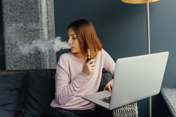 Vaping. Beautiful brunette smoke electronic cigarette in restaurant and working on her laptop. Vapor concept. Electronic cigarette with working