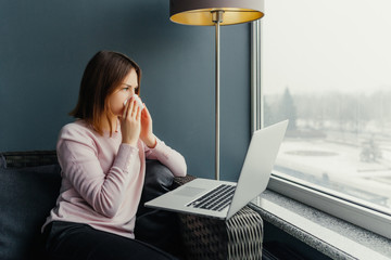 Young business woman blows her nose while working on laptop at cafe. Sick young girl is sitting cafe with laptop near the window
