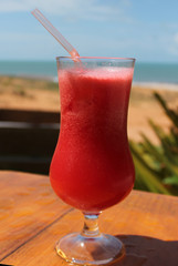 Copo de suco de melancia em uma mesa de madeira com praia ao fundo. Refresque-se com a fruta tropical!