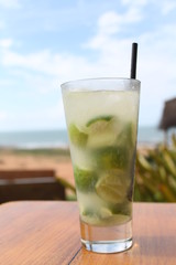 Copo de caipirinha de limão servido em copo de drink, sobre uma mesa de madeira, com uma praia tropical ao fundo, capturando o clima descontraído e tropical do Brasil