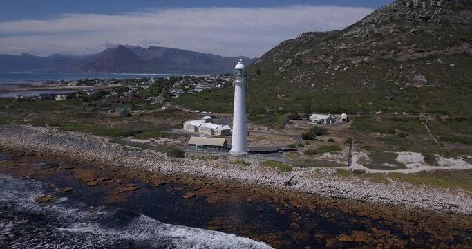 4K high quality aerial video sunny day view of historical Slangkop lighthouse built in 1914 and Kommetjie town in the background near beach at sea shore in Western Cape near Cape Town, South Africa