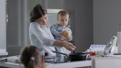 Mother making scrambled eggs, holding baby and talking on the phone when another child is jumping near her - Powered by Adobe