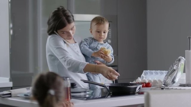 Mother making scrambled eggs, holding baby and talking on the phone when another child is jumping near her