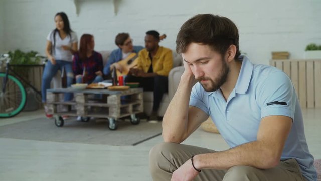 Young student guy feels upset and isolated while his friends celebrating party at home indoors