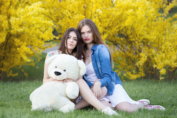 Two beautiful young women in park, sitting on grass, relaxing and sharing a teddy bear. Spring and summer lifestyle concept

