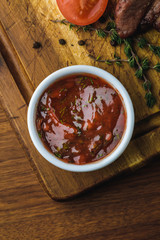 top view of bbq sauce in bowl and delicious roasted meat with tomato on wooden board