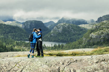 Portrait of two young people in love standing on plateau in Preikestolen, green forest and clouds on background. Woman in blue jacket. Norway.