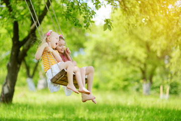 Two cute little sisters having fun on a swing together in beautiful summer garden on warm and sunny...