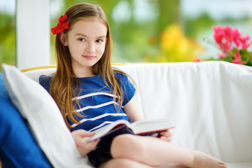 Adorable little girl reading a book in white living room on beautiful summer day.