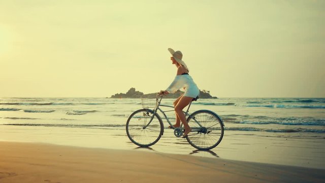 Woman in white and hat mooving on her bicycle along beach sand at summer time under burning sunset sky, lifestyle carefree.