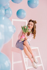 girl in a blue dress in a pink room with a bouquet of tulips in her hands sits on a ladder